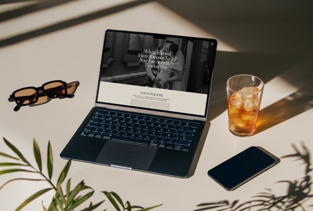 A laptop with modern homepage design of a website displayed on the screen with light cast from a window creating shadows. A pair of orange lensed glasses, an iced beverage, and a darken iphone scattered on either side of the laptop.