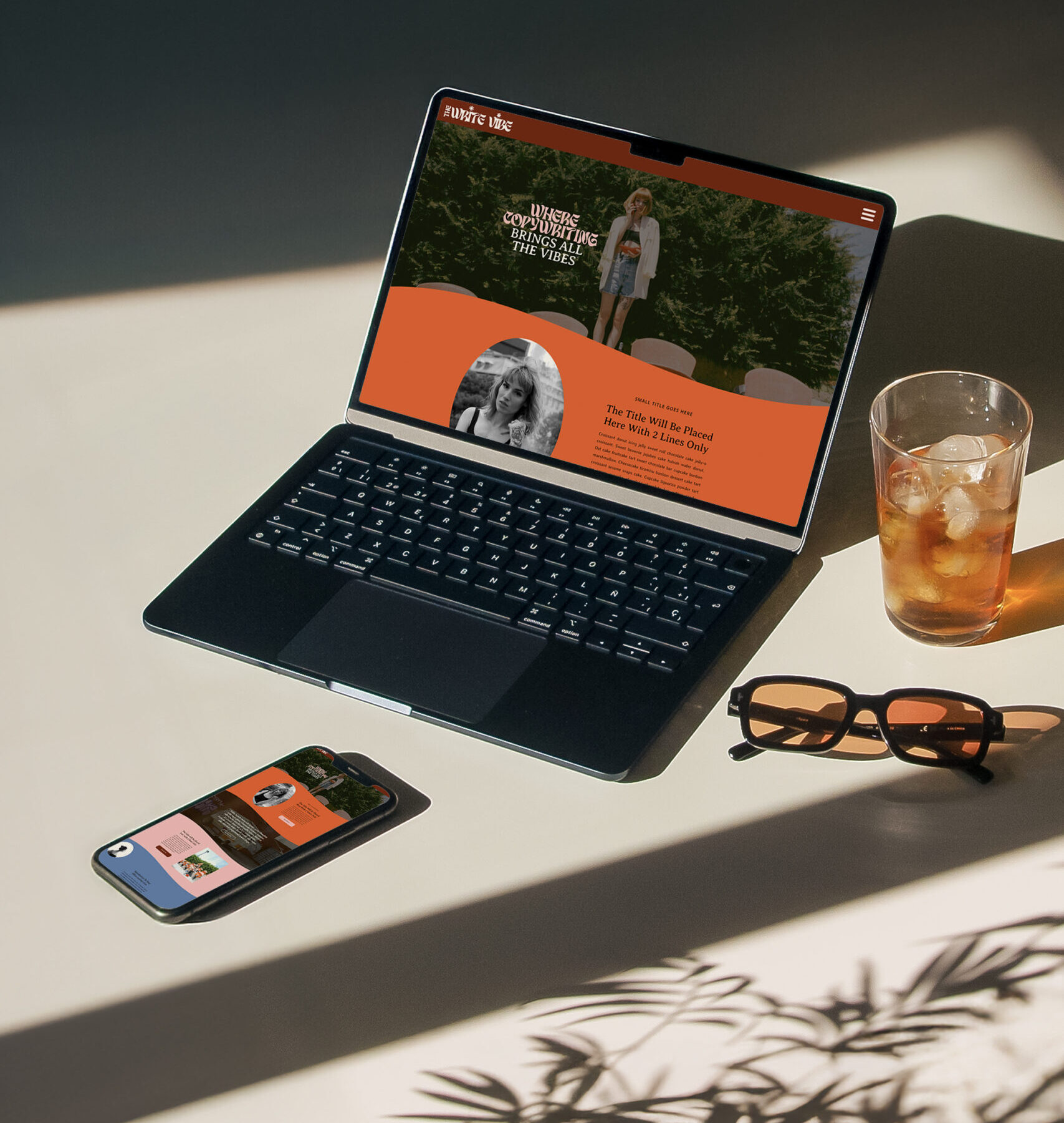 A laptop with fun, funky, colorful website homepage for a copywriter displayed on the screen with light cast from a window creating shadows. A pair of orange lensed glasses, an iced beverage, and a darken iphone scattered on either side of the laptop.
