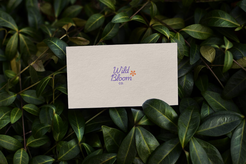 A cream colored business care with a purple, script text that reads Wild Bloom co with an orange flower detail lies in deep green colored, leafy foliage
